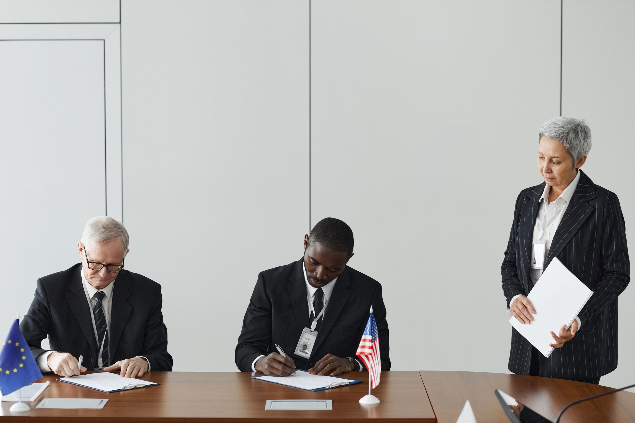 international-agreement-signing-eu-us-flags Executives signing an international agreement with EU and US flags displayed on a wooden table